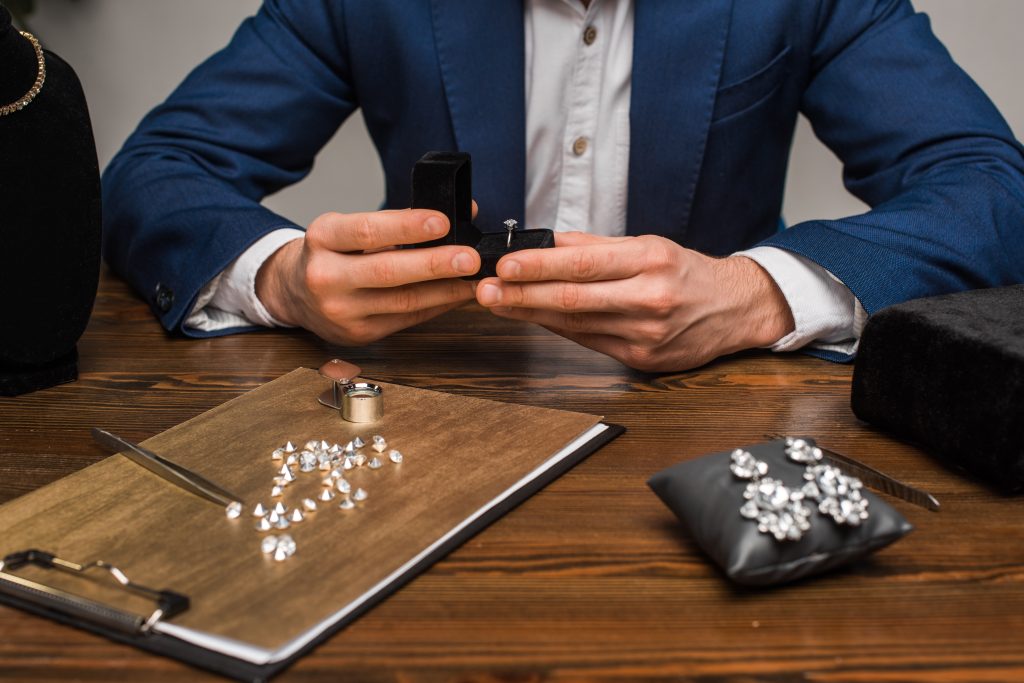 A jeweler sits at a table with earrings, a ring box, a loupe, and a spread of diamonds. 